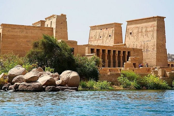 Malerischer Blick vom Wasser auf den Philae-Tempelkomplex, mit Felsen und grüner Vegetation.